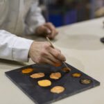 Hand painting on a black palette with various earthy colors, wearing a white sleeve, in an indoor workspace with blurred objects in the background.